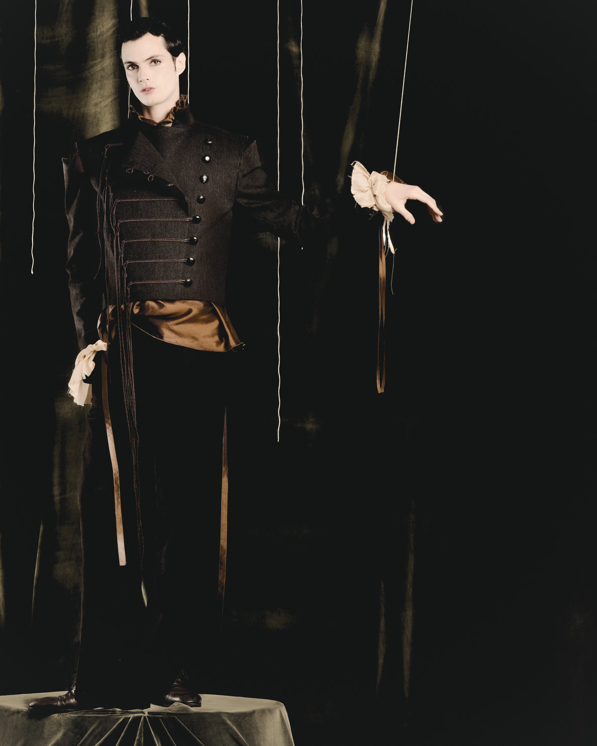 Adelina Bernard SS25 Pulling Strings campaign man in a dark master jacket with strings, hanging by puppet strings on a stage.