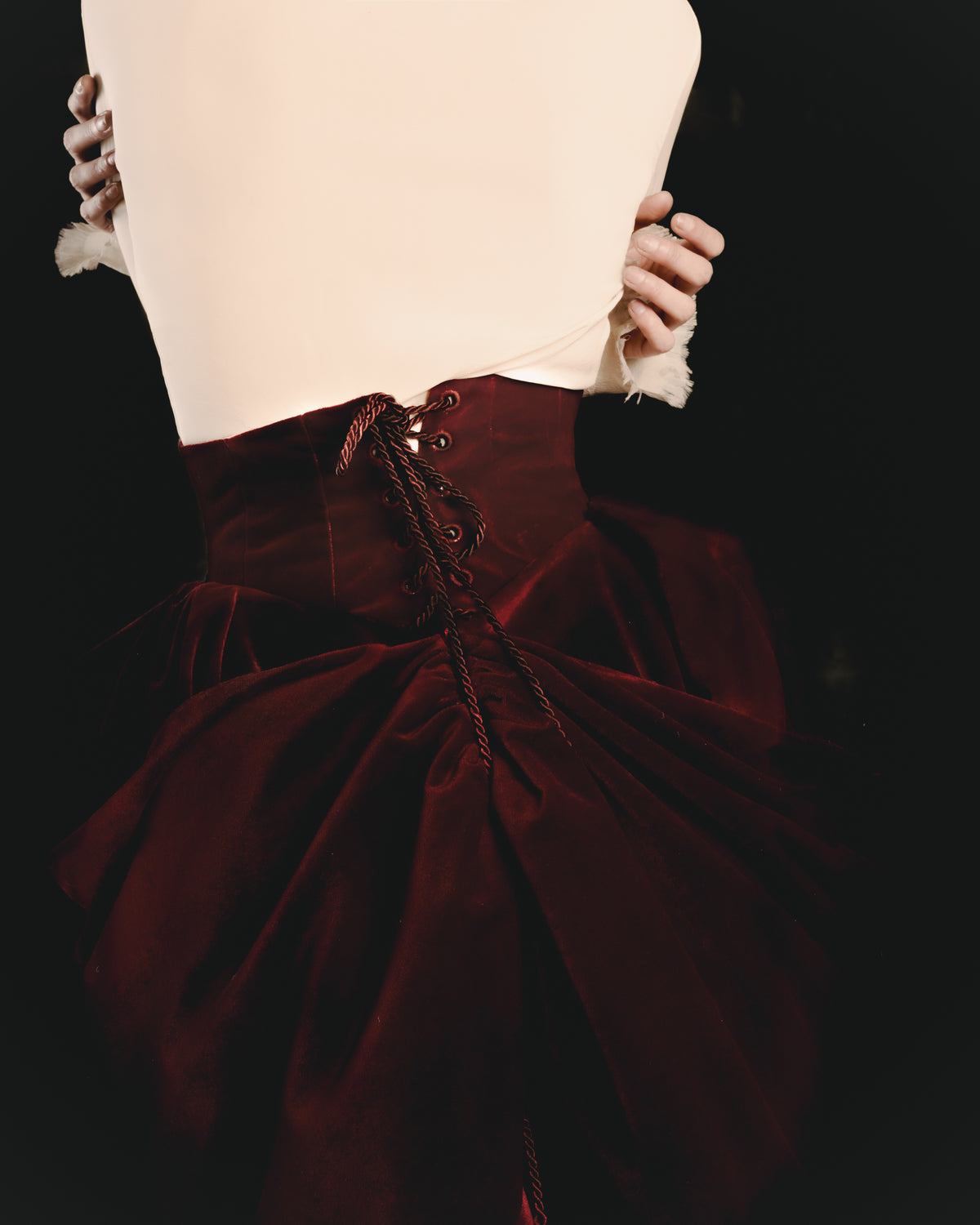 Adelina Bernard SS25 Pulling Strings campaign close-up of red velvet skirt and lace-up white shirt with hands wrapped around waist