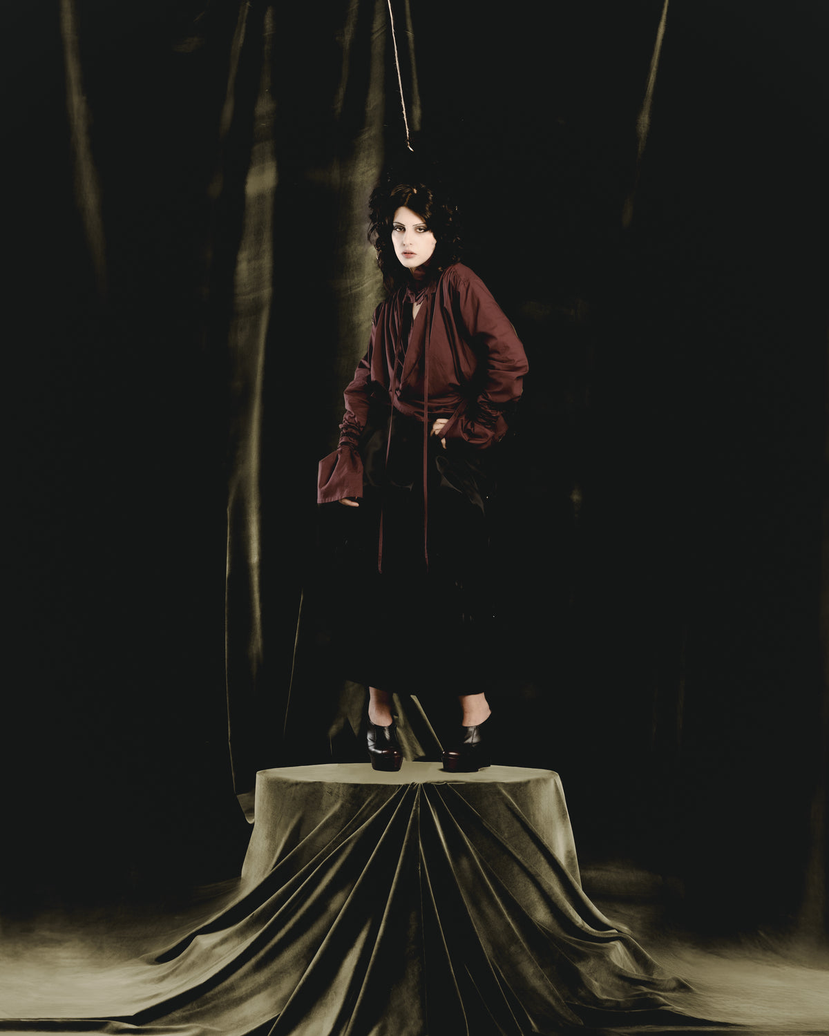 Adelina Bernard SS25 Pulling Strings campaign woman in aubergine blouse and black skirt standing on pedestal with hair suspended like puppet strings