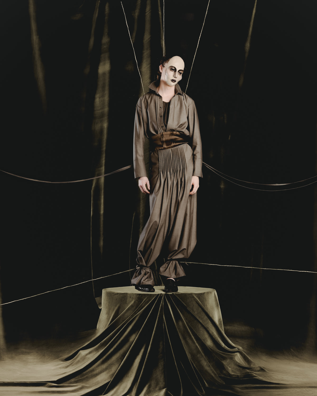 Adelina Bernard SS25 Pulling Strings campaign figure wearing a mask in grey pleated shirt and pants standing on pedestal with limbs  suspended like puppet strings
