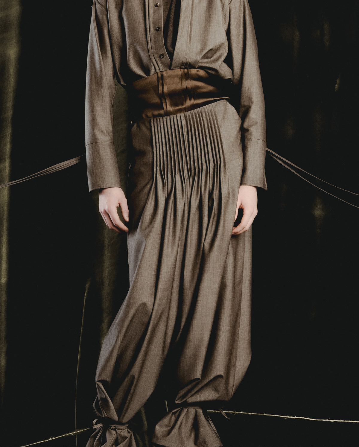 Adelina Bernard SS25 Pulling Strings campaign figure wearing grey pleated shirt and pants standing on pedestal with limbs  suspended like puppet strings