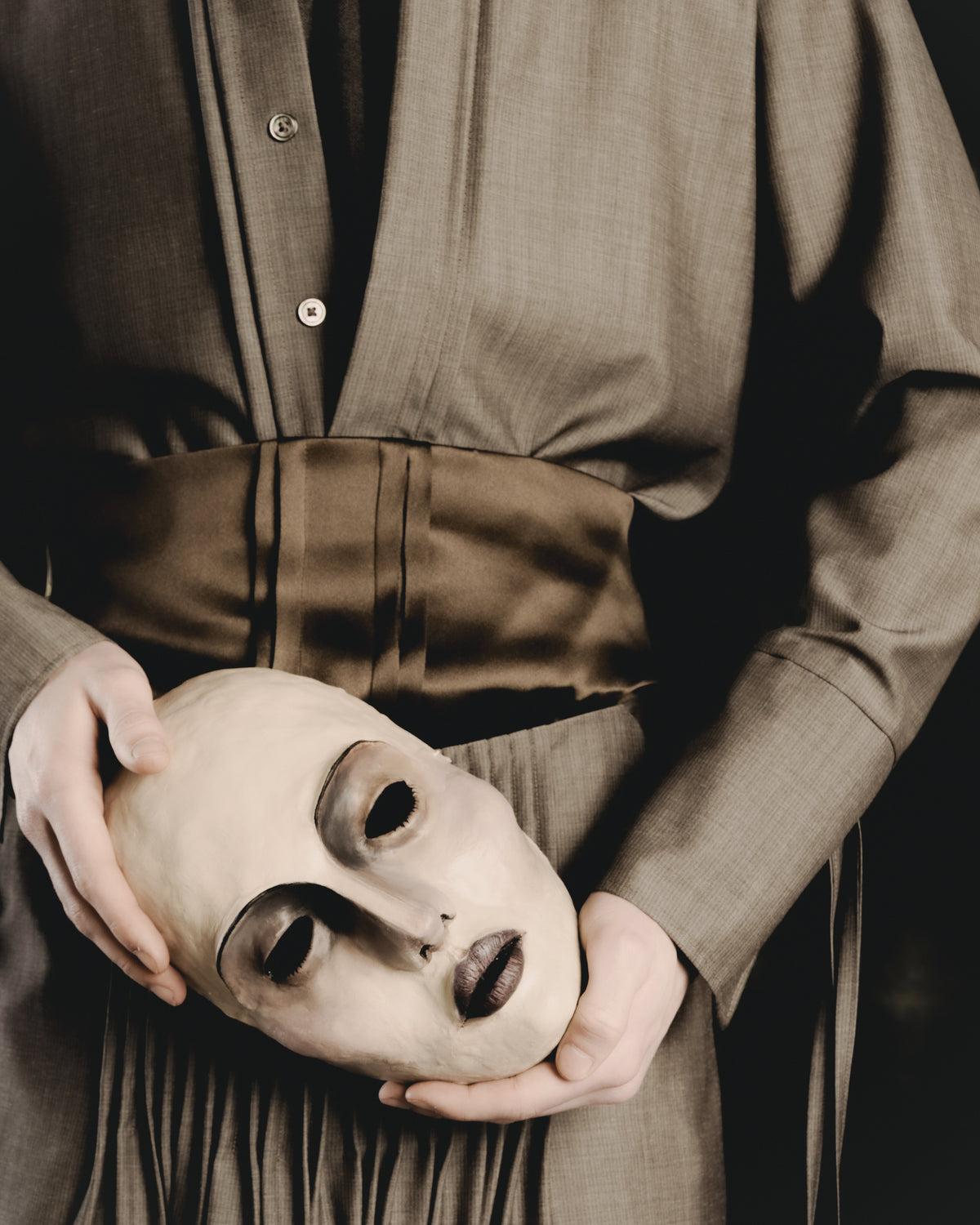 Adelina Bernard SS25 Pulling Strings campaign figure holding a mask wearing grey pleated shirt and pants with bronze waistband 