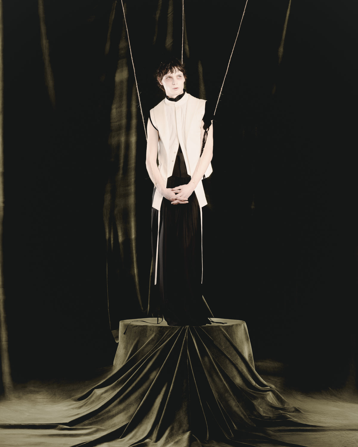 Adelina Bernard SS25 Pulling Strings campaign man in white layered vest and black trousers standing on pedestal with arms suspended in puppet strings